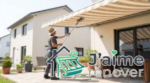 Un professionnel avec un chapeau nettoie un store banne rayé avec une brosse à long manche sur une terrasse ensoleillée.